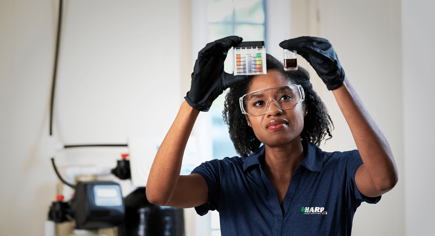 A Harp technician in a blue uniform and safety gear compares a water sample to a testing chart in front of a water softener system.