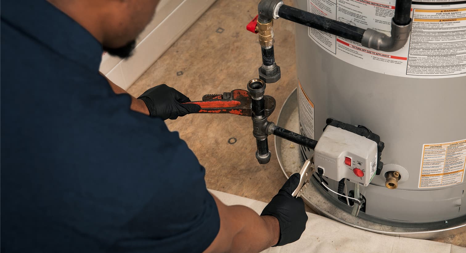 A Harp plumber wearing black gloves uses a pipe wrench to repair the gas line and control valve assembly on a residential water heater tank.